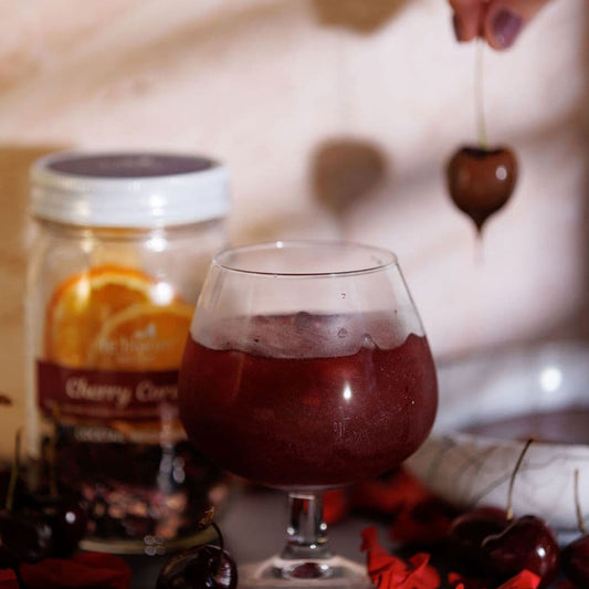 Glass of Cherry Cordial cocktail beside a Cherry Cordial infusion kit jar with fresh cherries and a hand lowering a cherry garnish into the drink.