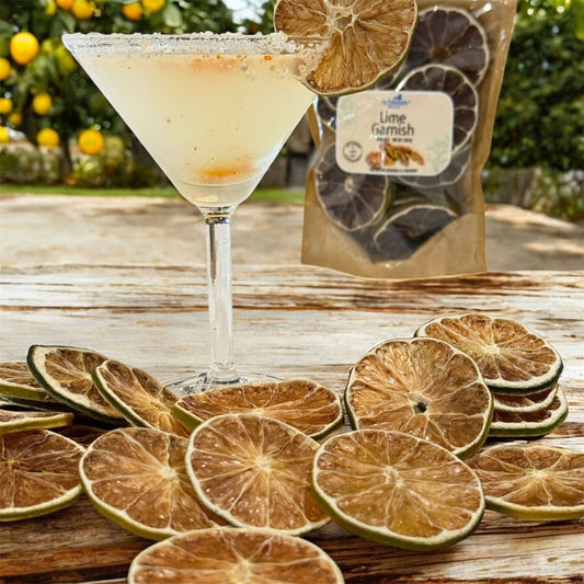 Dried lime slices displayed on a rustic wooden table, with a martini glass filled with a pale citrus drink and a bag labeled 'Lime Garnish' in the background.