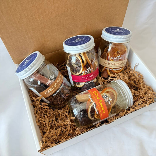 A gift box containing four glass jars of dried botanicals and spices arranged on brown crinkle paper inside a white box with a brown lid. The jars include Espresso Martini, Cosmo, Old Fashioned, and Citrus Sangria.