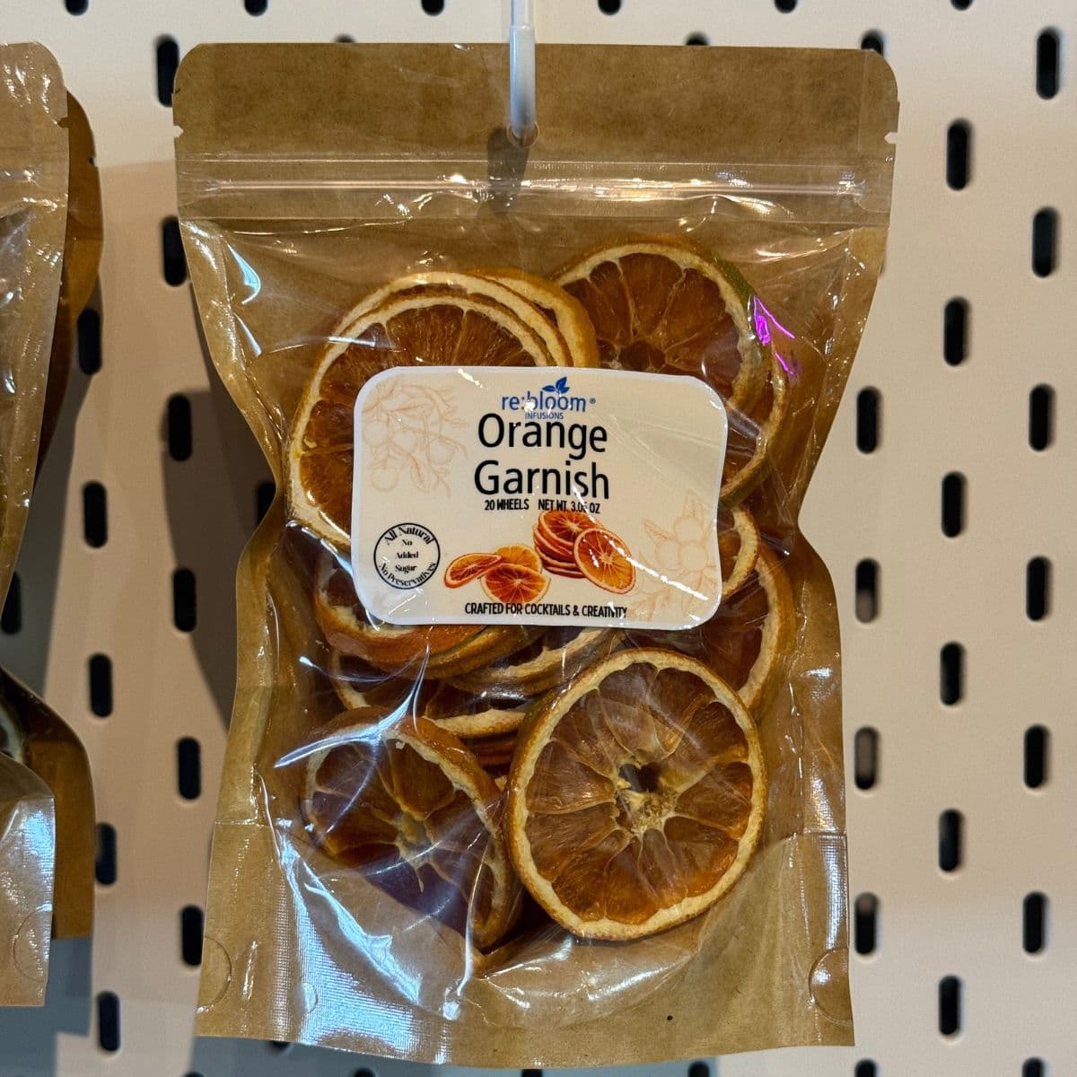A clear plastic pouch of Re:Imagined Orange Garnish on a beige pegboard background. The packaging is brown kraft paper with a white label showing the brand name and product name, with dried orange slices visible inside.