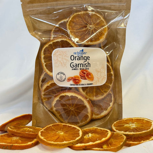 A clear resealable pouch labeled 're:blooms Orange Garnish' filled with dried orange slices, with additional dried orange slices scattered in front on a white background.