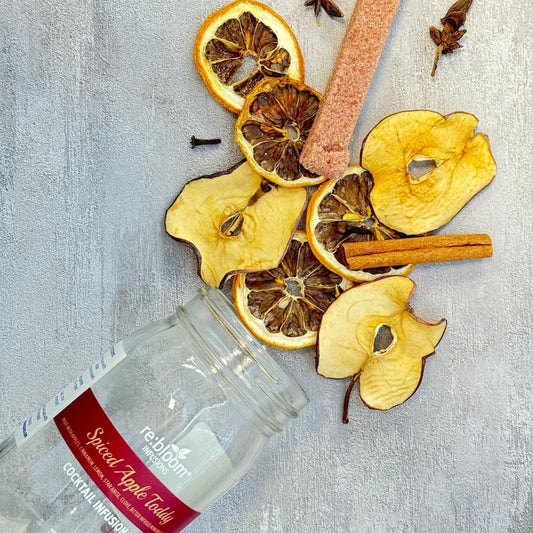 Spiced apple toddy ingredients including dried apples, oranges, and cinnamon sticks on a textured surface with a jar labeled 're: bloom'.