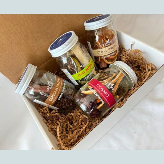 A gift box containing four glass jars of dried botanicals and spices nestled in brown paper shred. The jars feature labels for 'Espresso Martini', 'Old Fashion', 'Spicy Margarita', and 'Cosmo'.
