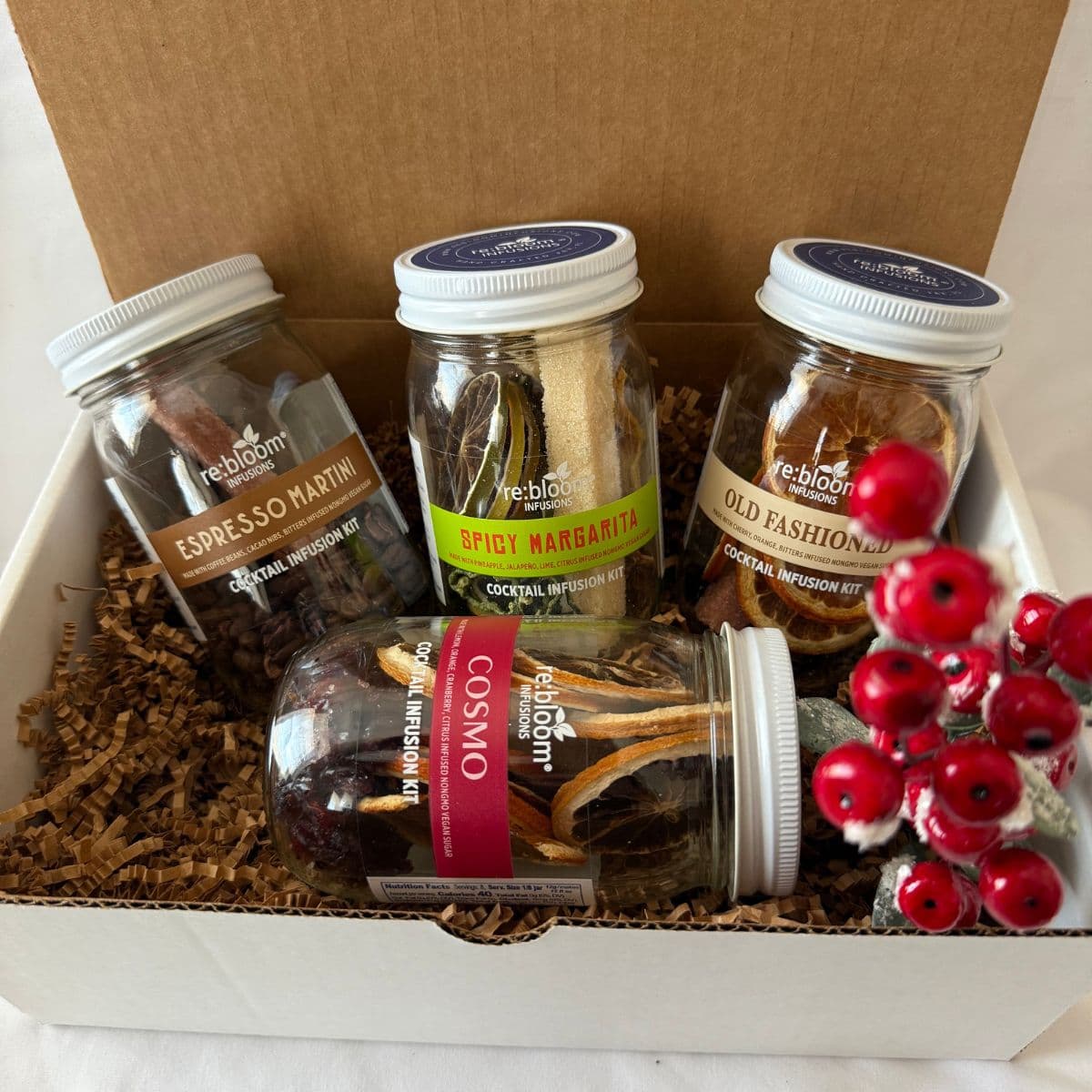 A gift box containing four glass jars of Reblooms cocktail mixtures, arranged in brown crinkle paper filler with decorative red berries in the front. The jars are labeled Espresso Martini, Spicy Margarita, Old Fashioned, and Cosmo.