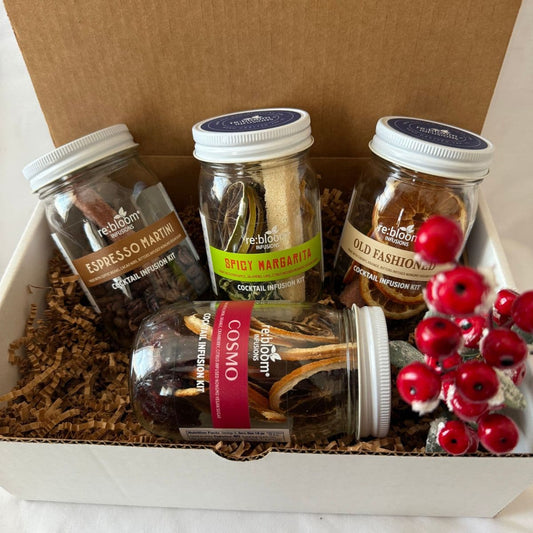 A gift box containing four glass jars of Reblooms cocktail mixtures, arranged in brown crinkle paper filler with decorative red berries in the front. The jars are labeled Espresso Martini, Spicy Margarita, Old Fashioned, and Cosmo.