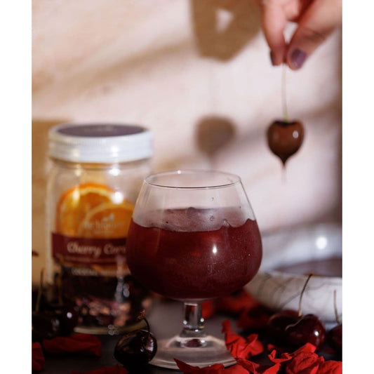 Glass of Cherry Cordial cocktail beside a Cherry Cordial infusion kit jar with fresh cherries and a hand lowering a cherry garnish into the drink.