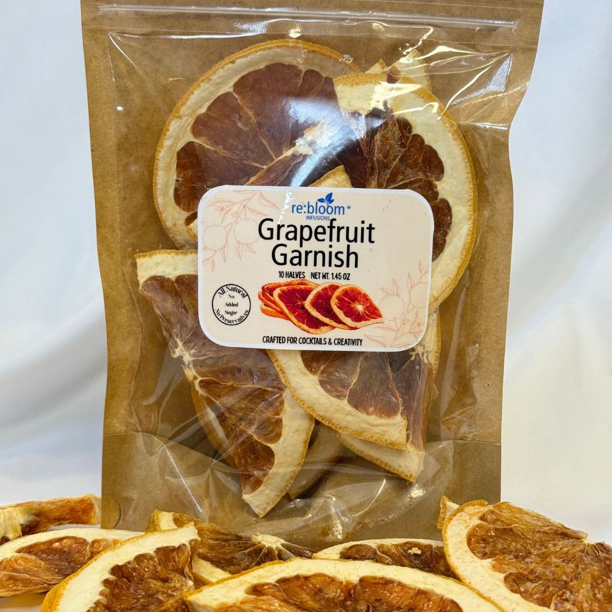 A clear plastic pouch of Rebloom Grapefruit Garnish on a white background, with dried grapefruit slices arranged in front. The label reads 'rebloom Grapefruit Garnish' and shows a white background with brown and red grapefruit imagery.