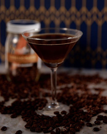 Cocktail in a martini glass surrounded by coffee beans with a blurred bottle in the background