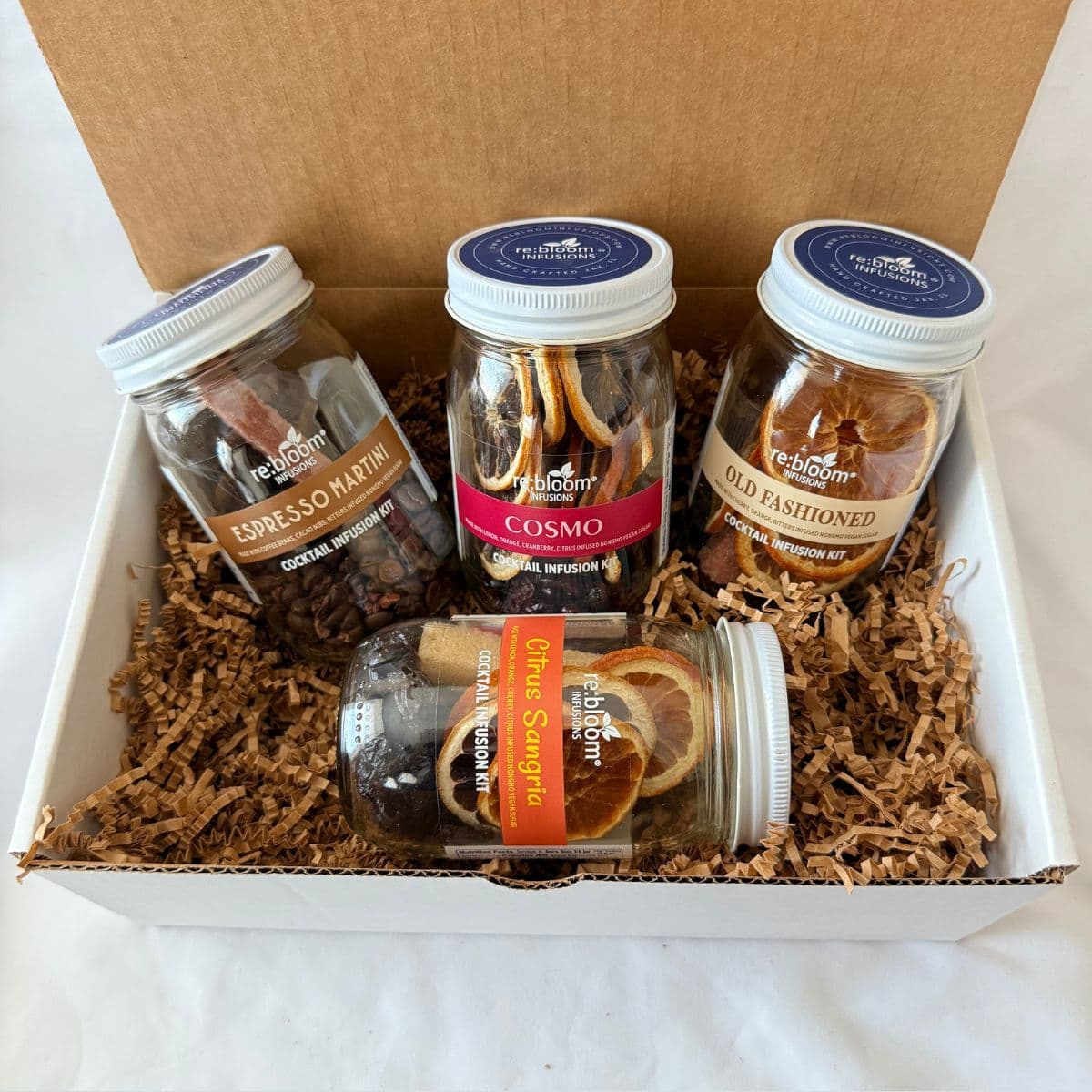 A gift box of four jars of Rebloom infusion jars, arranged in brown paper shred inside a white box with a brown cardboard lid. The jars include Espresso Martini, Cosmo, Old Fashioned, and Citrus Sangria.