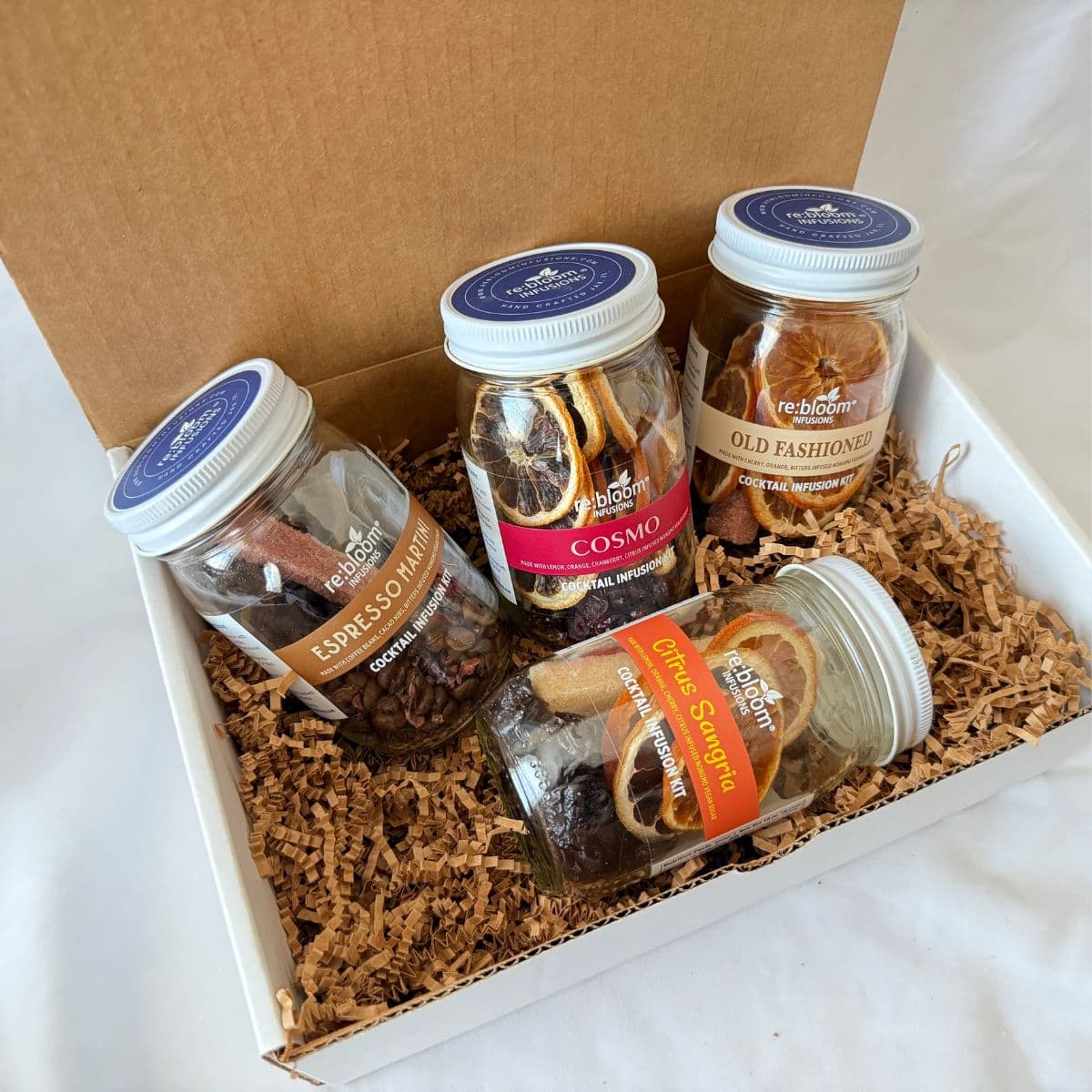 A gift box containing four glass jars of dried botanicals and spices arranged on brown crinkle paper inside a white box with a brown lid. The jars include Espresso Martini, Cosmo, Old Fashioned, and Citrus Sangria.