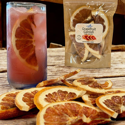 Glass of pink drink with grapefruit garnish, dried grapefruit slices, and a package of grapefruit garnish on a wooden surface.