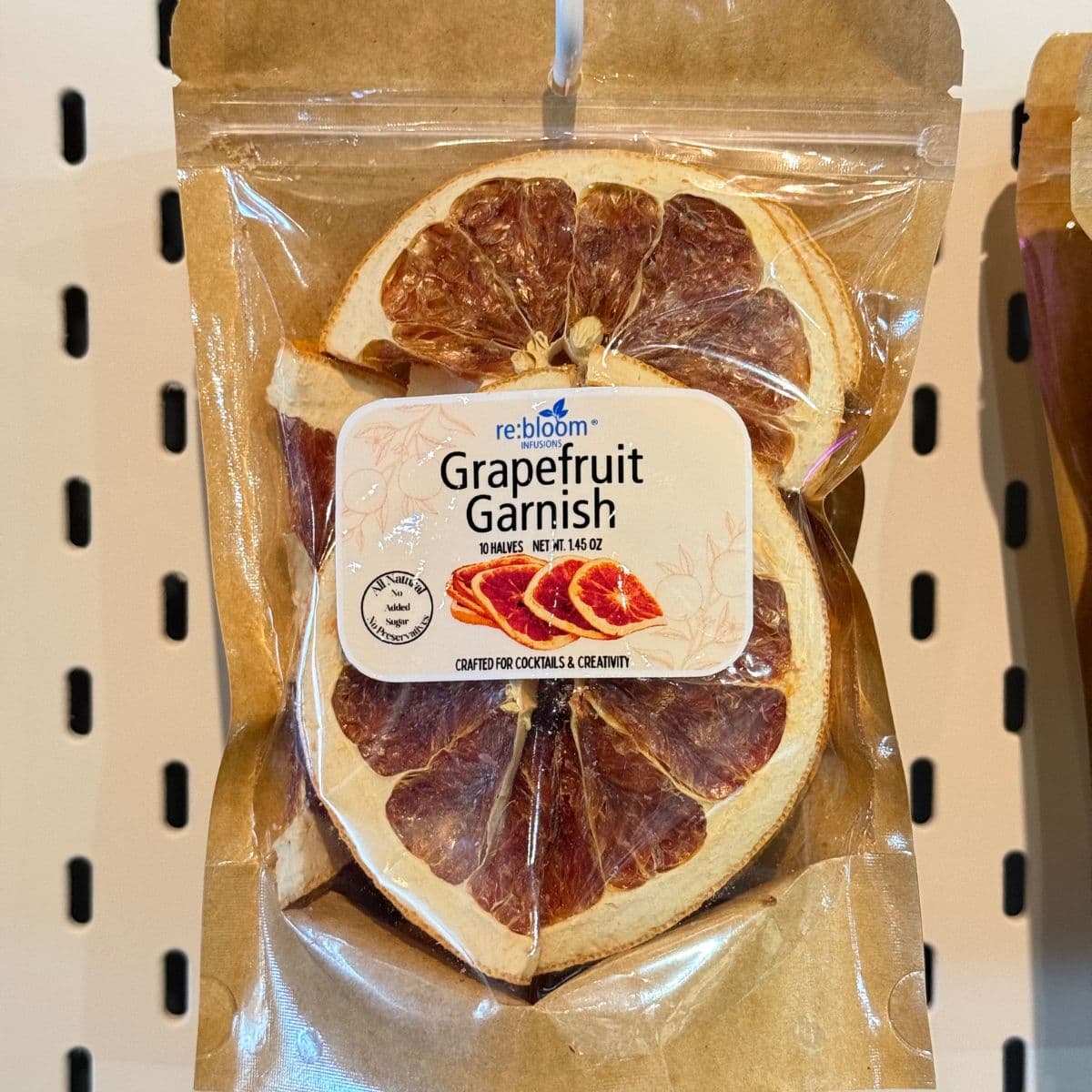 A package of Rebloom Grapefruit Garnish, featuring dried grapefruit slices inside a brown kraft paper bag with a clear plastic window and a white product label.