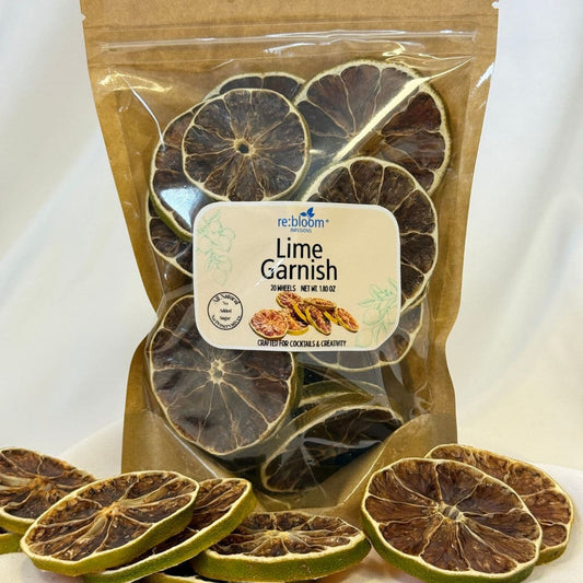 A clear stand-up pouch of Re:Bloom Lime Garnish dried citrus slices, with several slices scattered in front on a white surface against a white draped background.