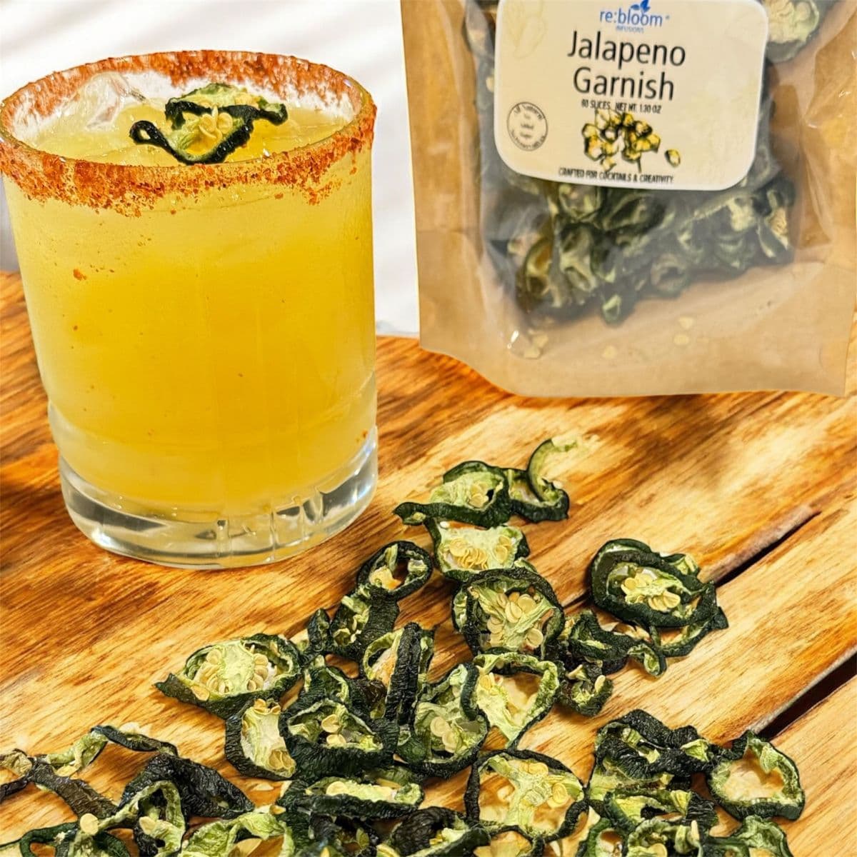 Spicy margarita with chili rim and dried jalapeño garnish beside packaged jalapeño cocktail garnish on a wooden surface.