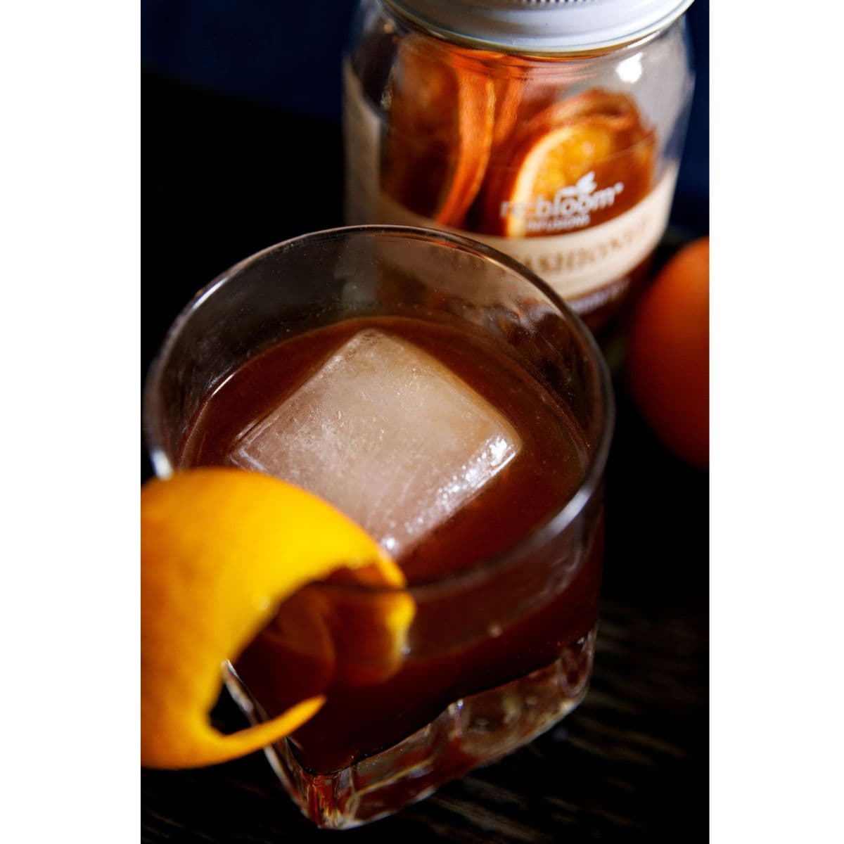 Glass of iced old fashioned drink with a orange twist next to a re:bloom old fashioned infusion jar.