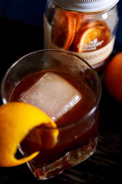 Glass of iced old fashioned drink with a orange twist next to a re:bloom old fashioned infusion jar.