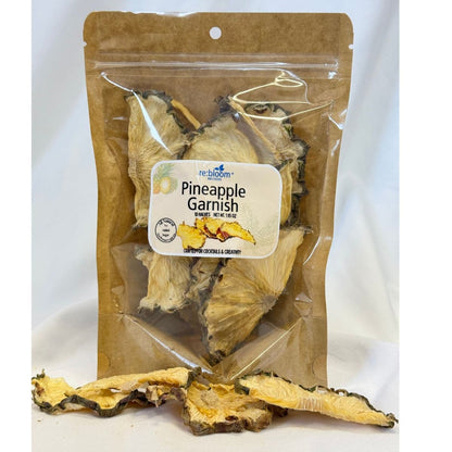 Packaged pineapple garnish slices with visible re:bloom Infusions label displayed on a white background with dried pineapple pieces in front.