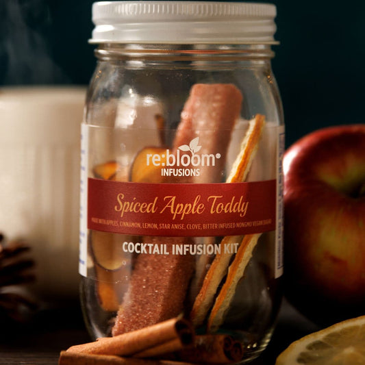 Jar of 'Spiced Apple Toddy' cocktail infusion kit with cinnamon sticks and apples on a dark background.
