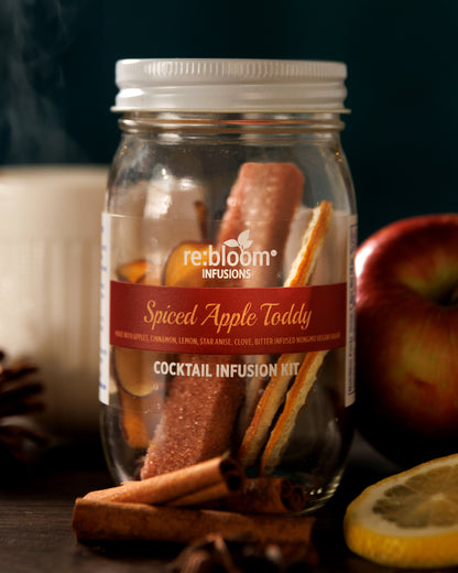 Spiced Apple Toddy cocktail infusion kit in a glass jar with cinnamon sticks and apples on a dark background.