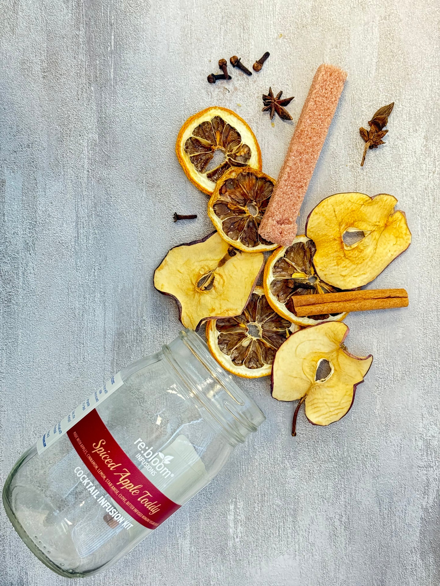 Spiced apple toddy ingredients including dried apples, cinnamon sticks, and a glass bottle on a gray surface.