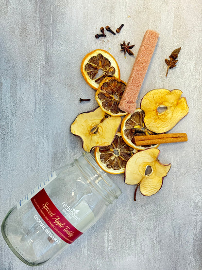Spiced apple toddy ingredients including dried apples, cinnamon sticks, and a glass bottle on a gray surface.