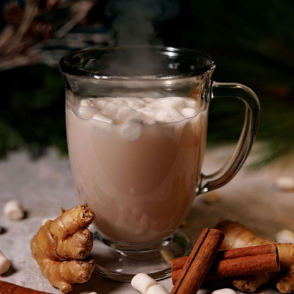 Glass mug filled with a hot beverage, likely chai, garnished with marshmallows, surrounded by spices on a rustic surface.
