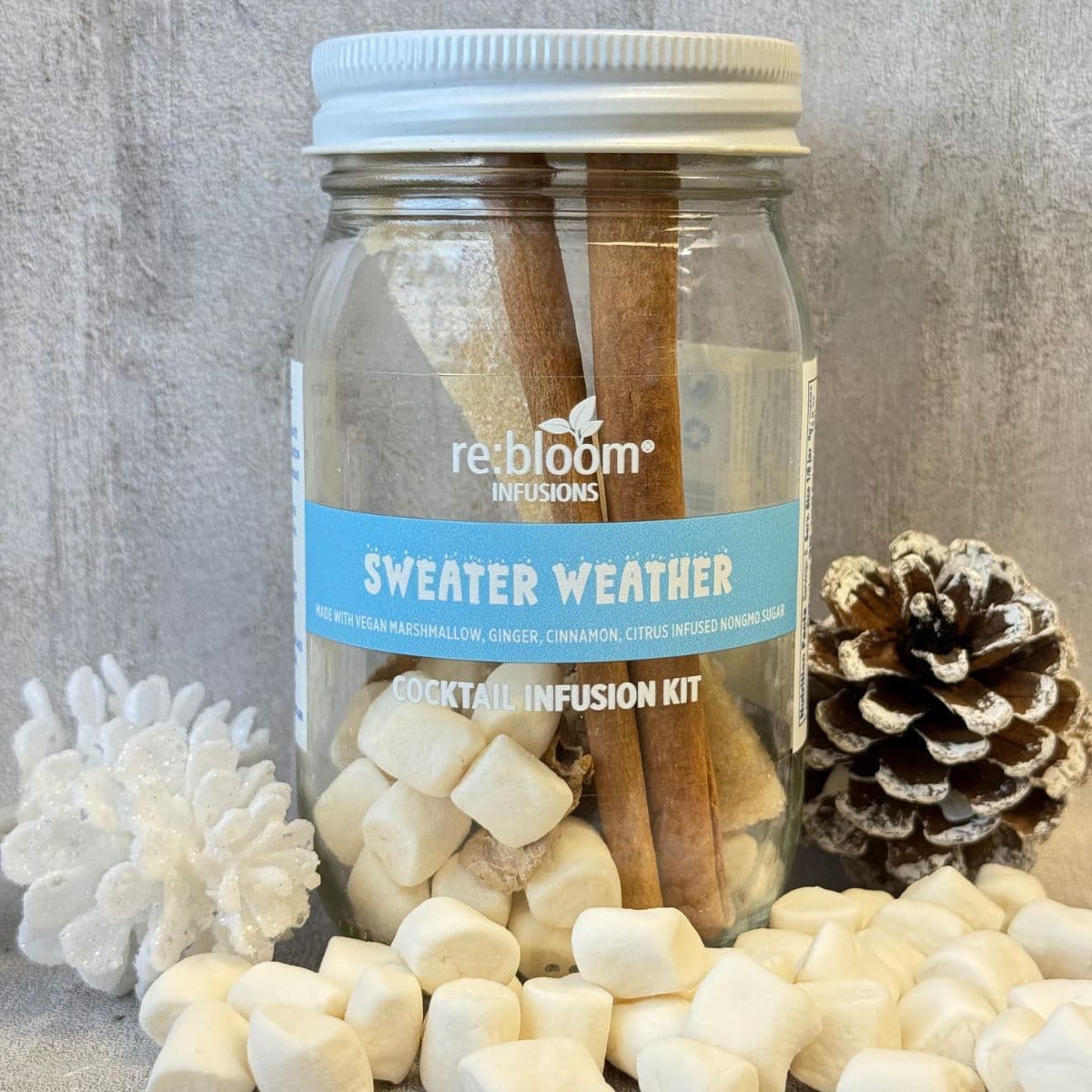 Cocktail infusion kit labeled 'Sweater Weather' by re:bloom with cinnamon sticks, marshmallows, and a pine cone on a textured surface.