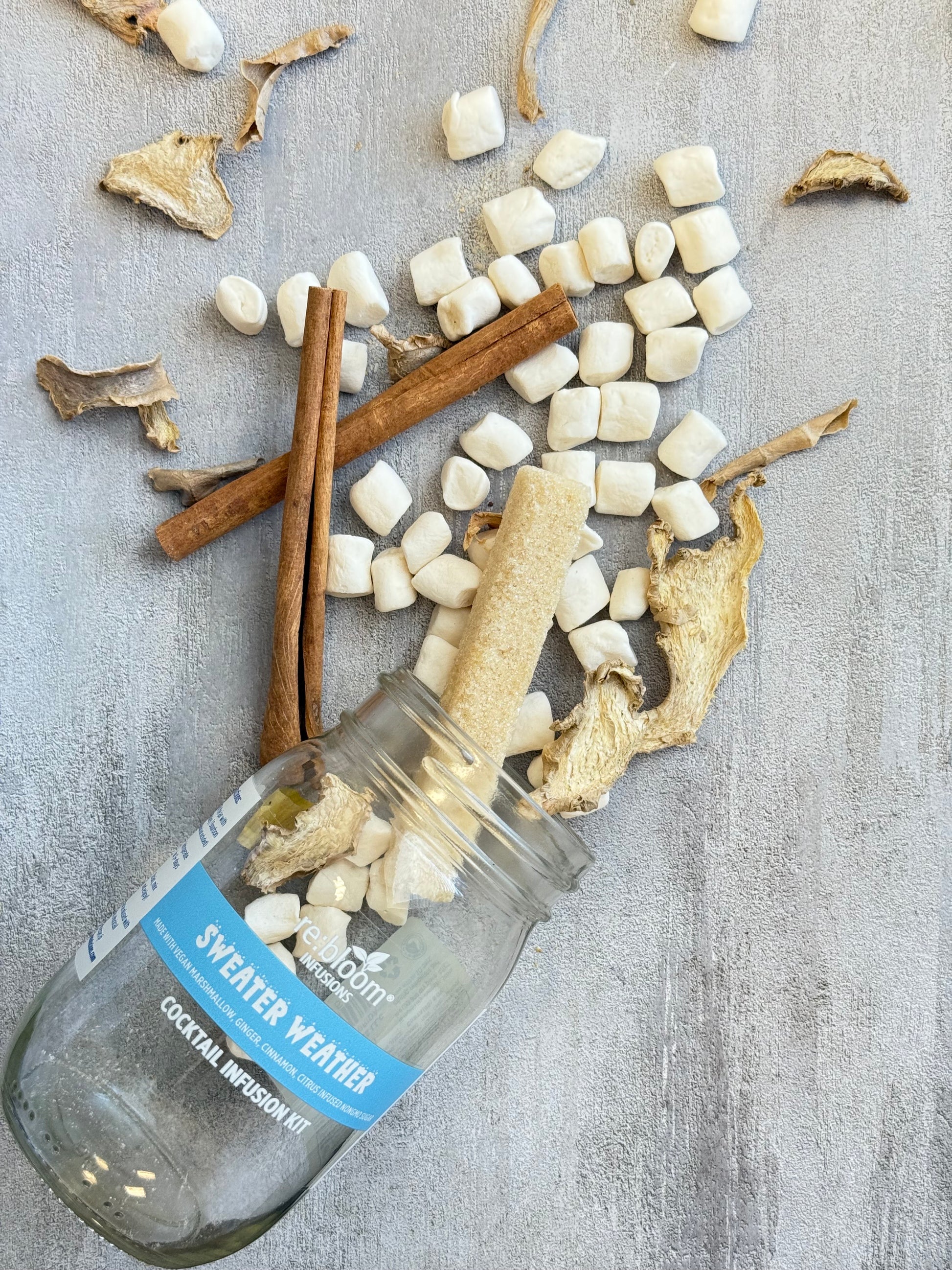 Glass bottle labeled 'Sweater Weather' with dried roots, marshmallows, and cinnamon sticks on a textured surface.