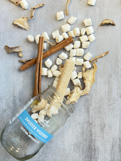 Glass bottle labeled 'Sweater Weather' with dried roots, marshmallows, and cinnamon sticks on a textured surface.
