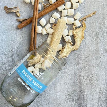 Glass bottle with 'Sweater Weather' cocktail infusion kit label, surrounded by cinnamon sticks and marshmallows on a textured surface.
