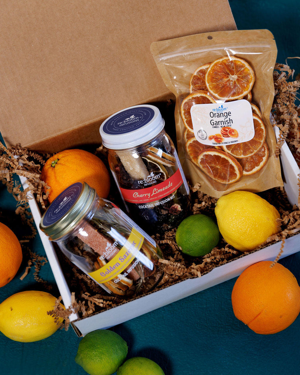 Gourmet gift box with cocktail infusion jars and a bag of orange garnish on a blue surface.